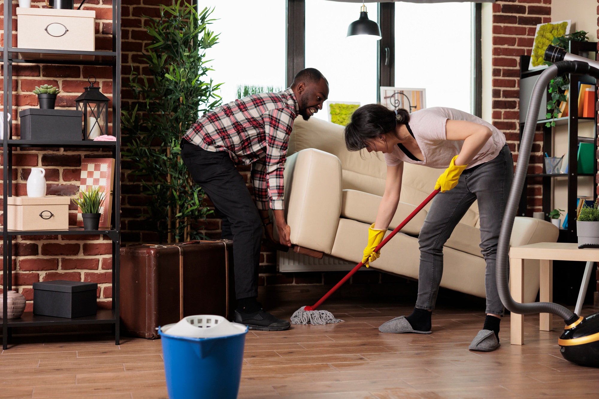 Multiracial couple cleaning their living room during spring cleaning, with one partner lifting the couch while the other mops the floor