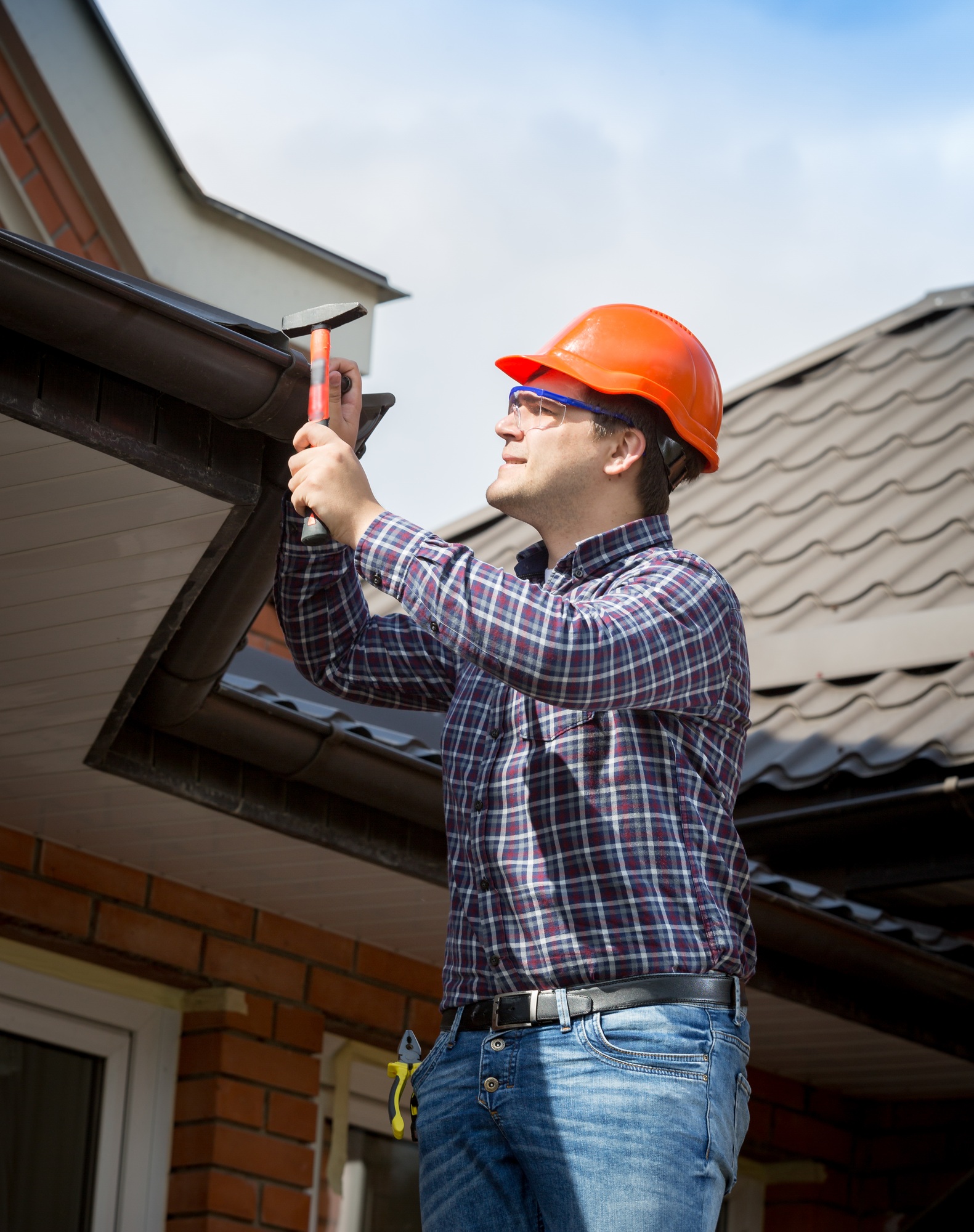 Young handyman using a hammer to secure nails into the roof shingles during a repair, with tools and roofing materials scattered nearby