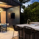 Contemporary outdoor kitchen with stainless steel appliances, built-in grill, and a sleek dining table with seating for eight, set on a stone patio surrounded by greenery.