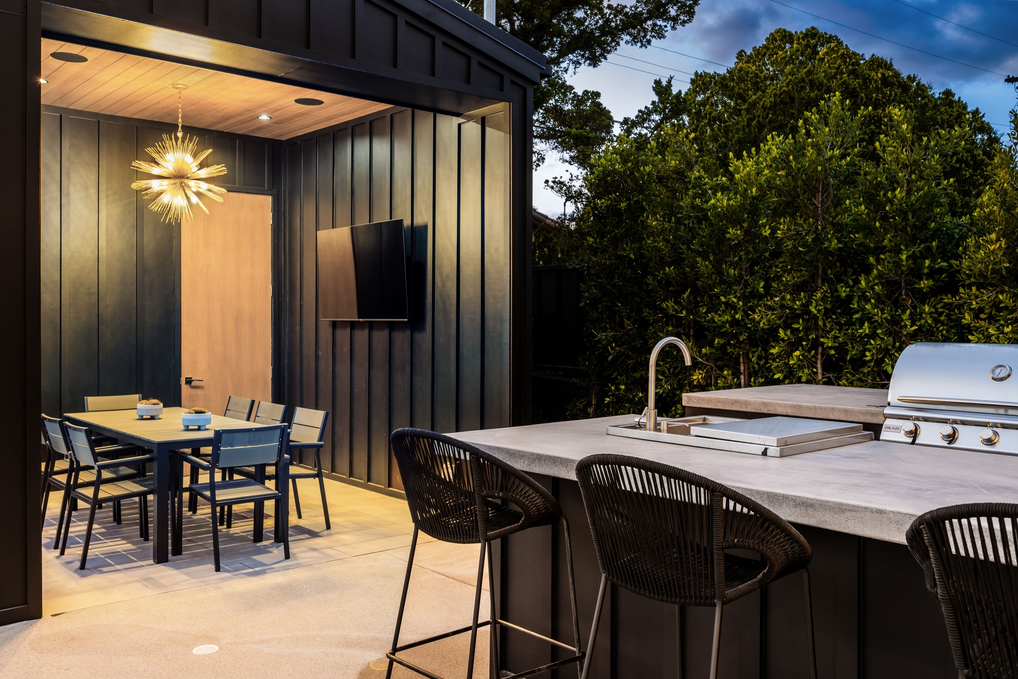 Contemporary outdoor kitchen with stainless steel appliances, built-in grill, and a sleek dining table with seating for eight, set on a stone patio surrounded by greenery.